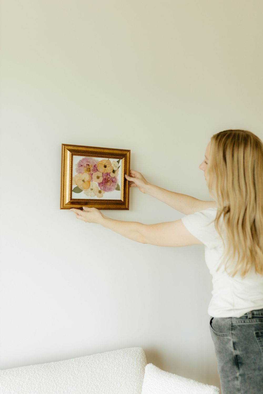 Blonde woman hanging a gold framed pressed flower artwork on a light gray wall.