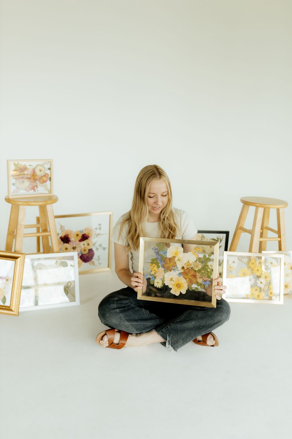 Blonde woman sitting cross-legged holding a large gold frame with pressed yellow flowers.
