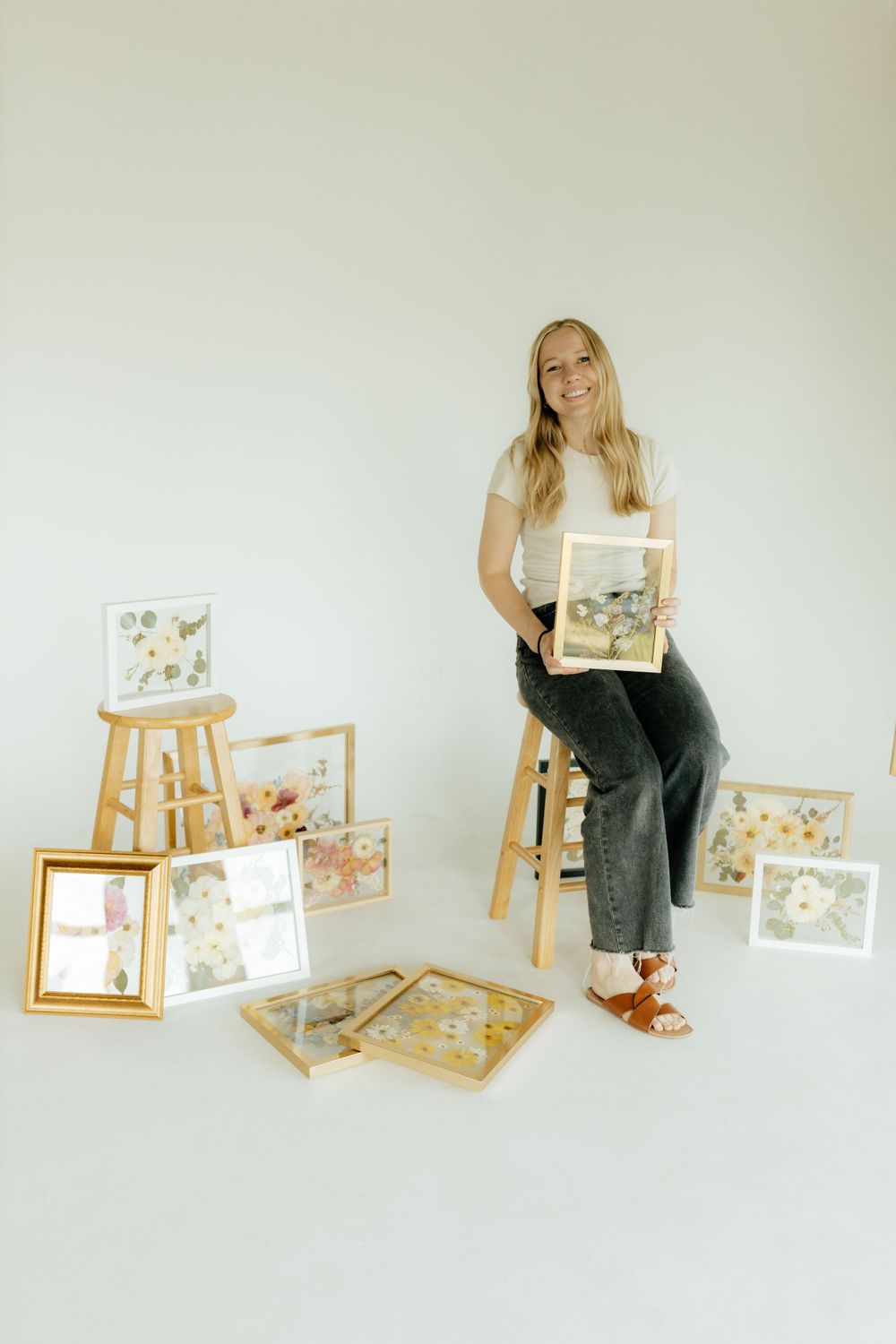 Smiling blonde woman sitting on stool surrounded by multiple pressed flower framed artworks.