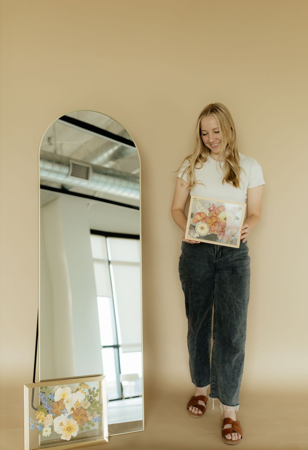 Smiling blonde woman holding pressed flower artwork beside a tall arched mirror in a studio.