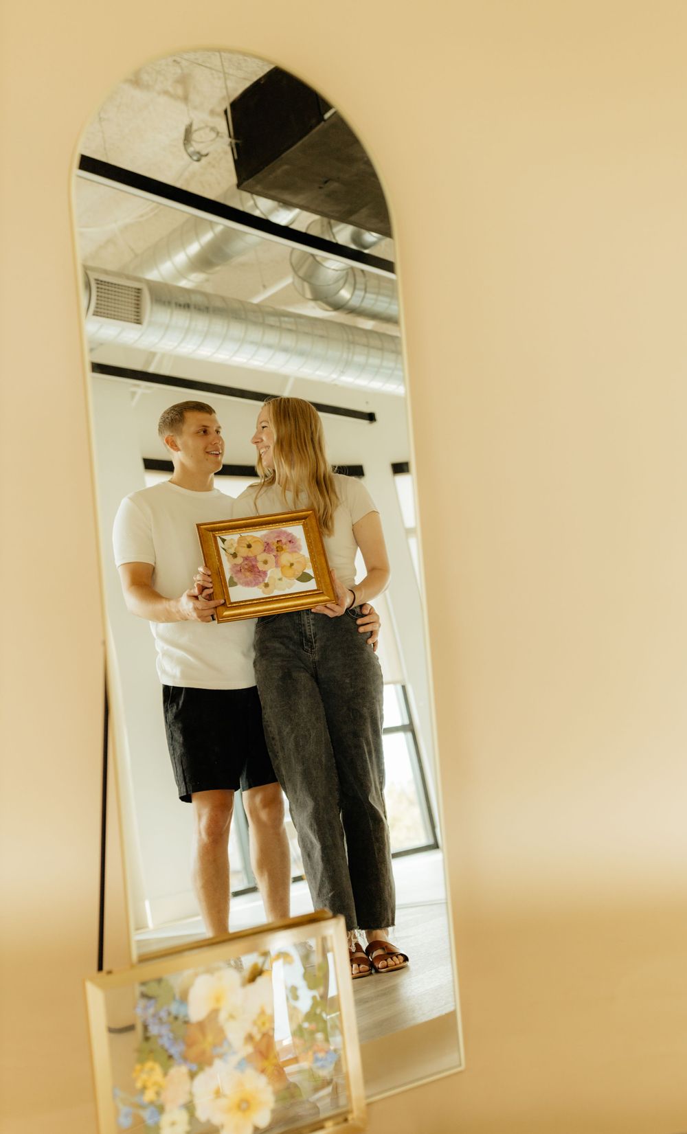 Couple smiling and holding framed pressed flower art reflected in a large arched floor mirror.