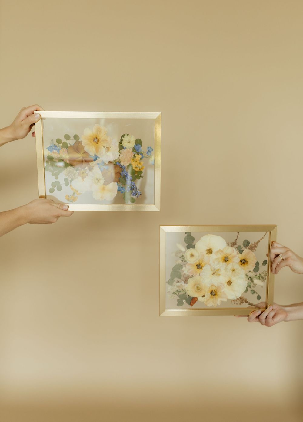Two hands holding gold-framed pressed flower artwork against a warm beige background.