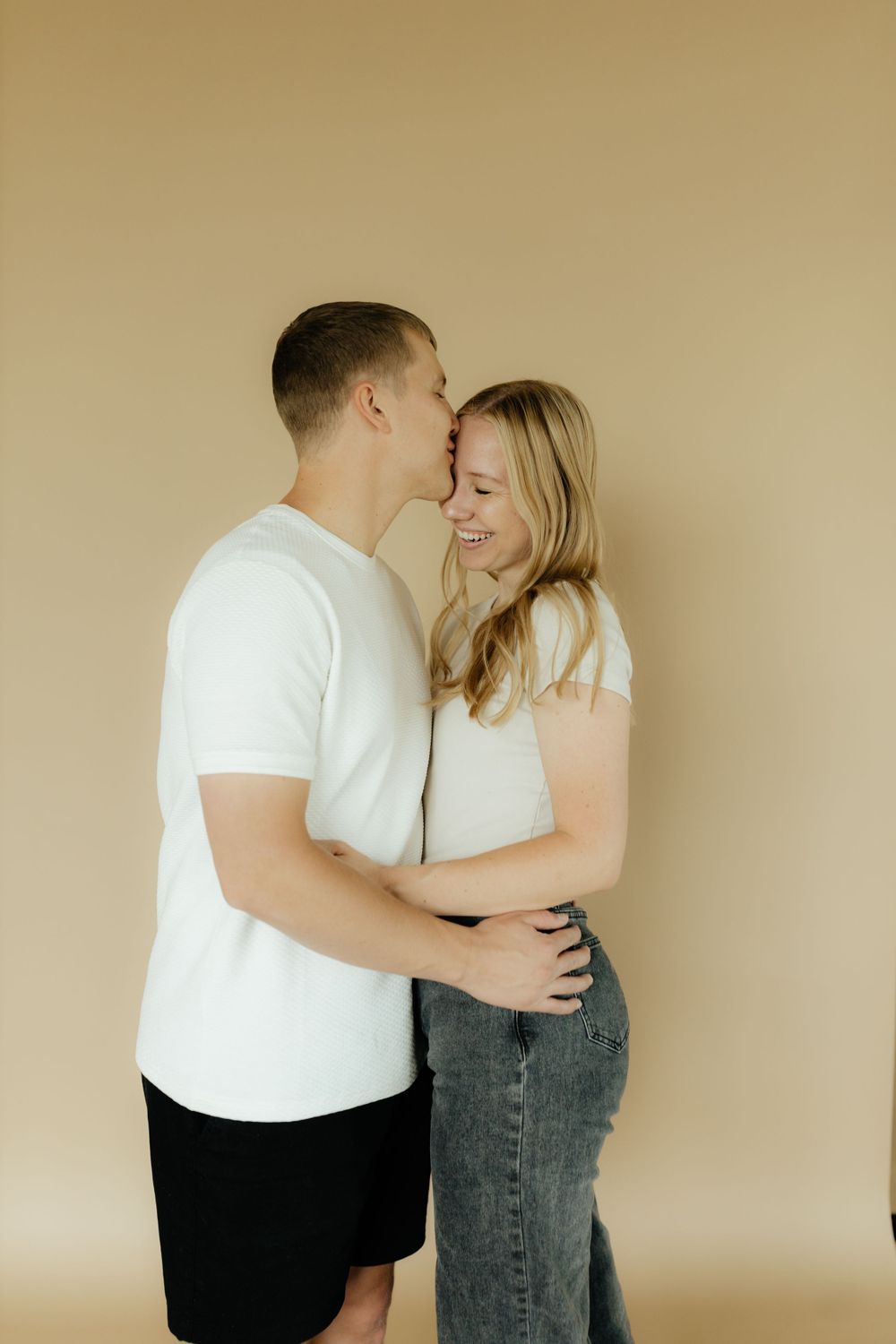 Happy couple embracing and sharing a kiss in a minimalist beige studio setting.