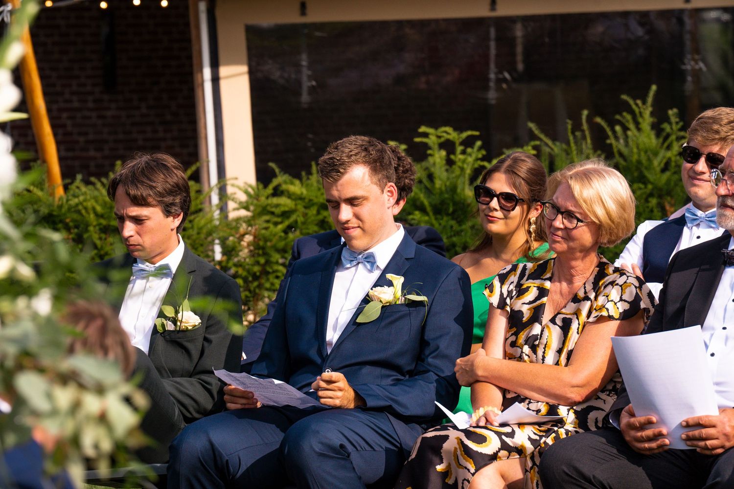 Wedding guests seated outdoors at formal ceremony with greenery and brick building backdrop.