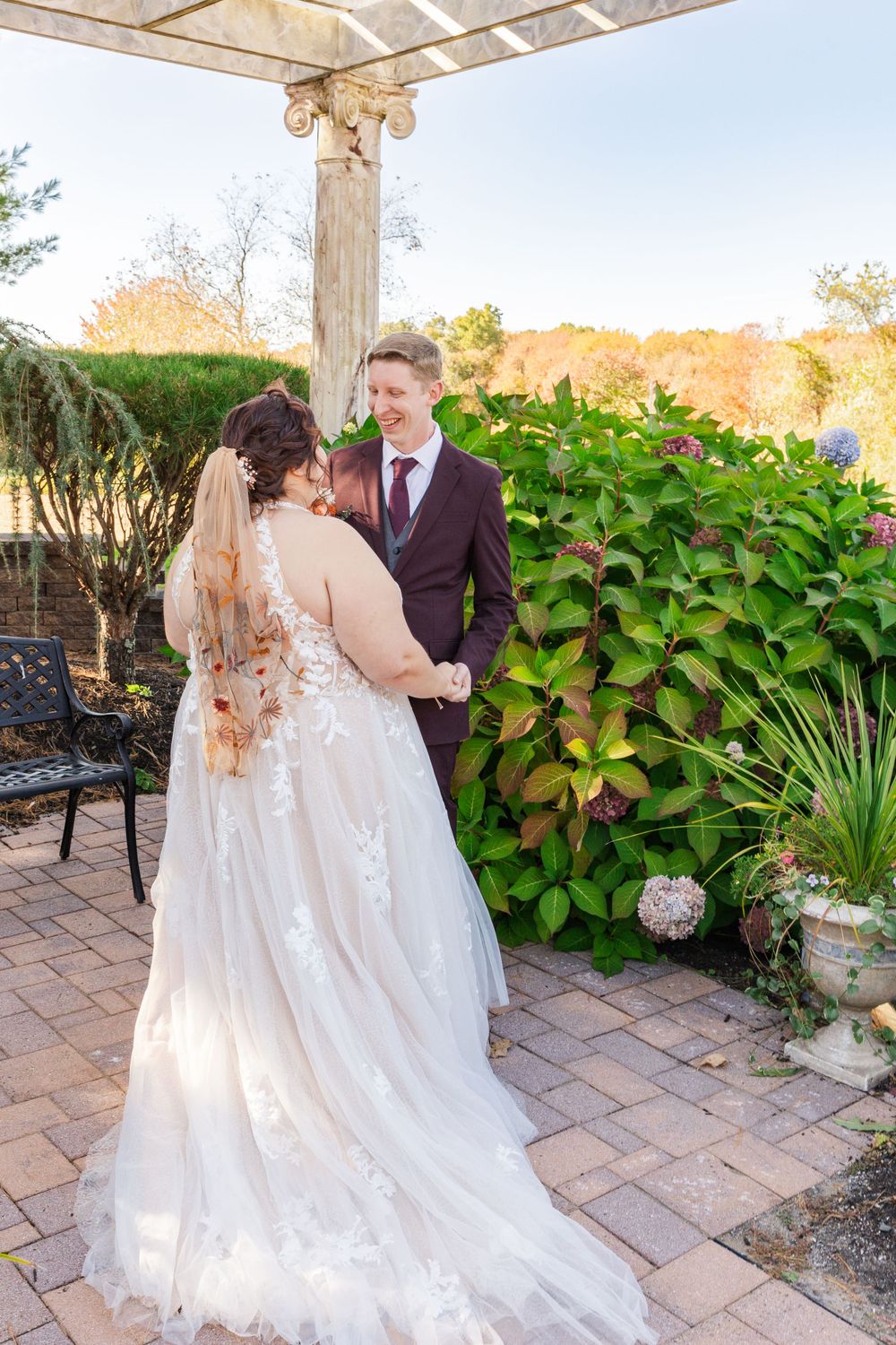 A couple poses under a white pergola in a lush garden setting with brick paving.
