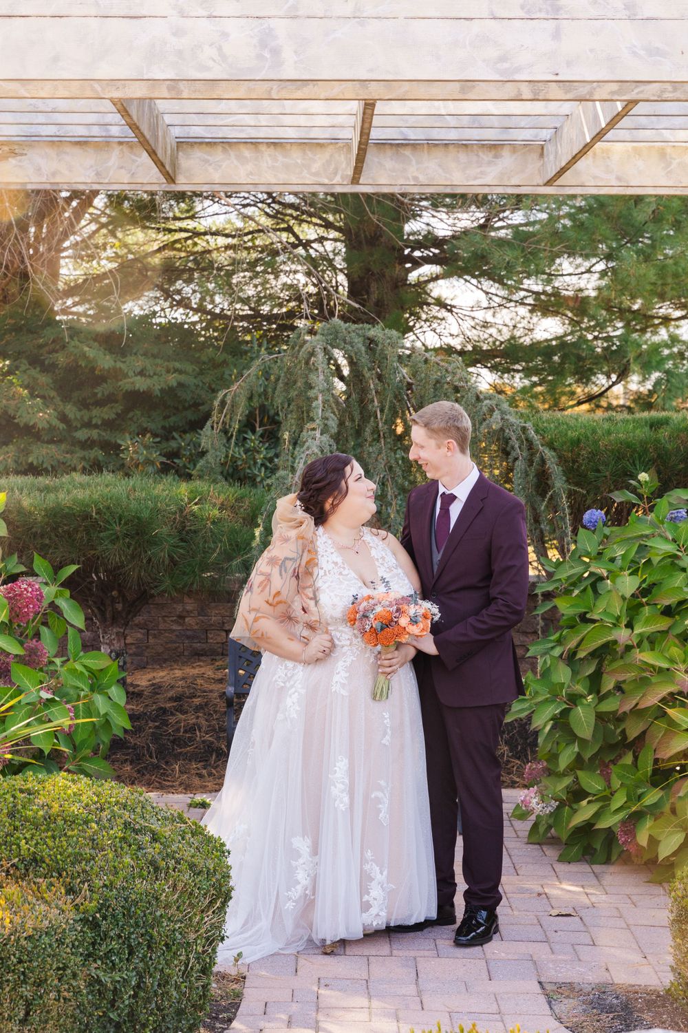 A couple stands under a pergola holding orange flowers in a manicured garden setting.