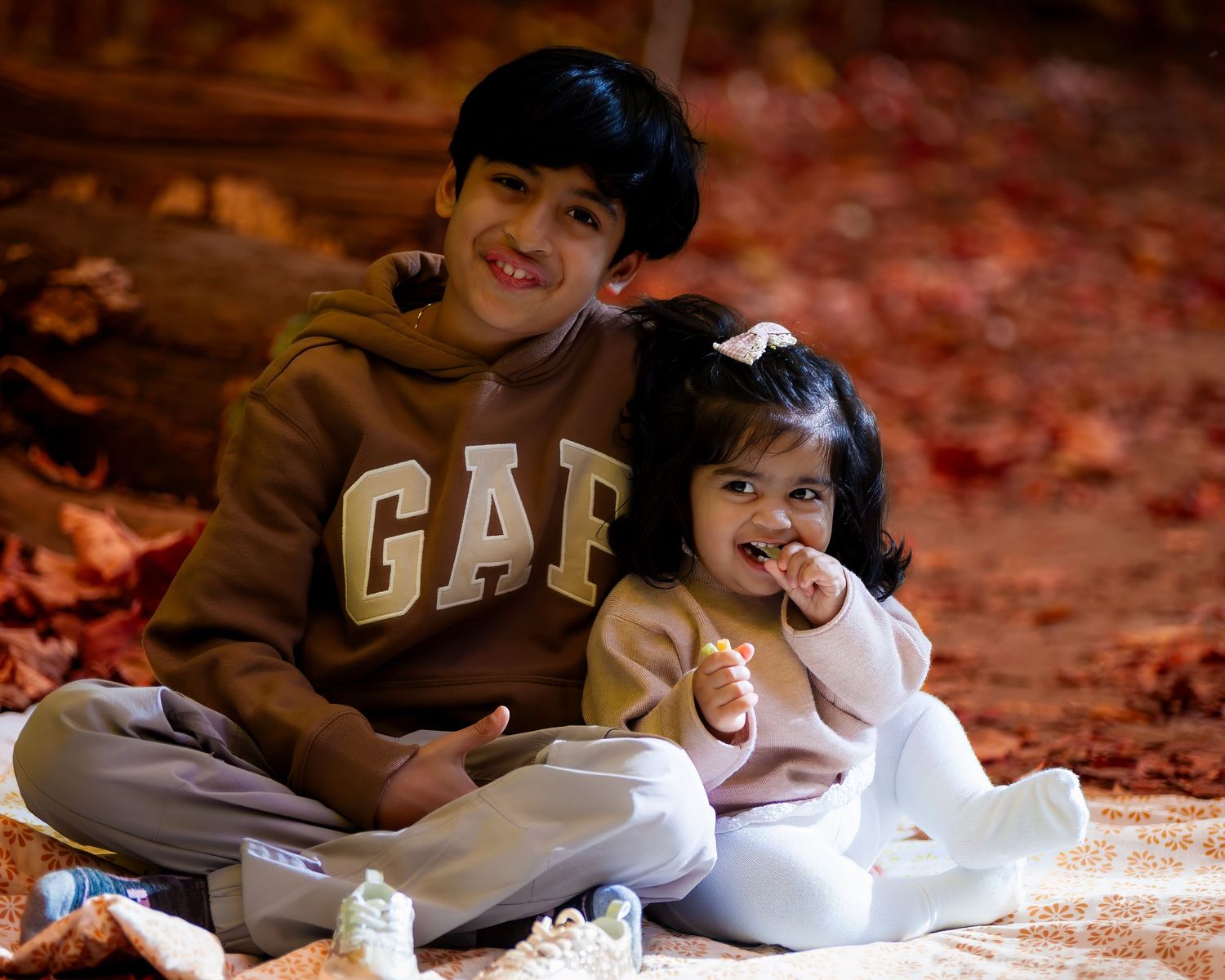 Elder brother and little sister enjoying a joyful moment together in an outdoor fall photoshoot, with the vibrant autumn leaves creating