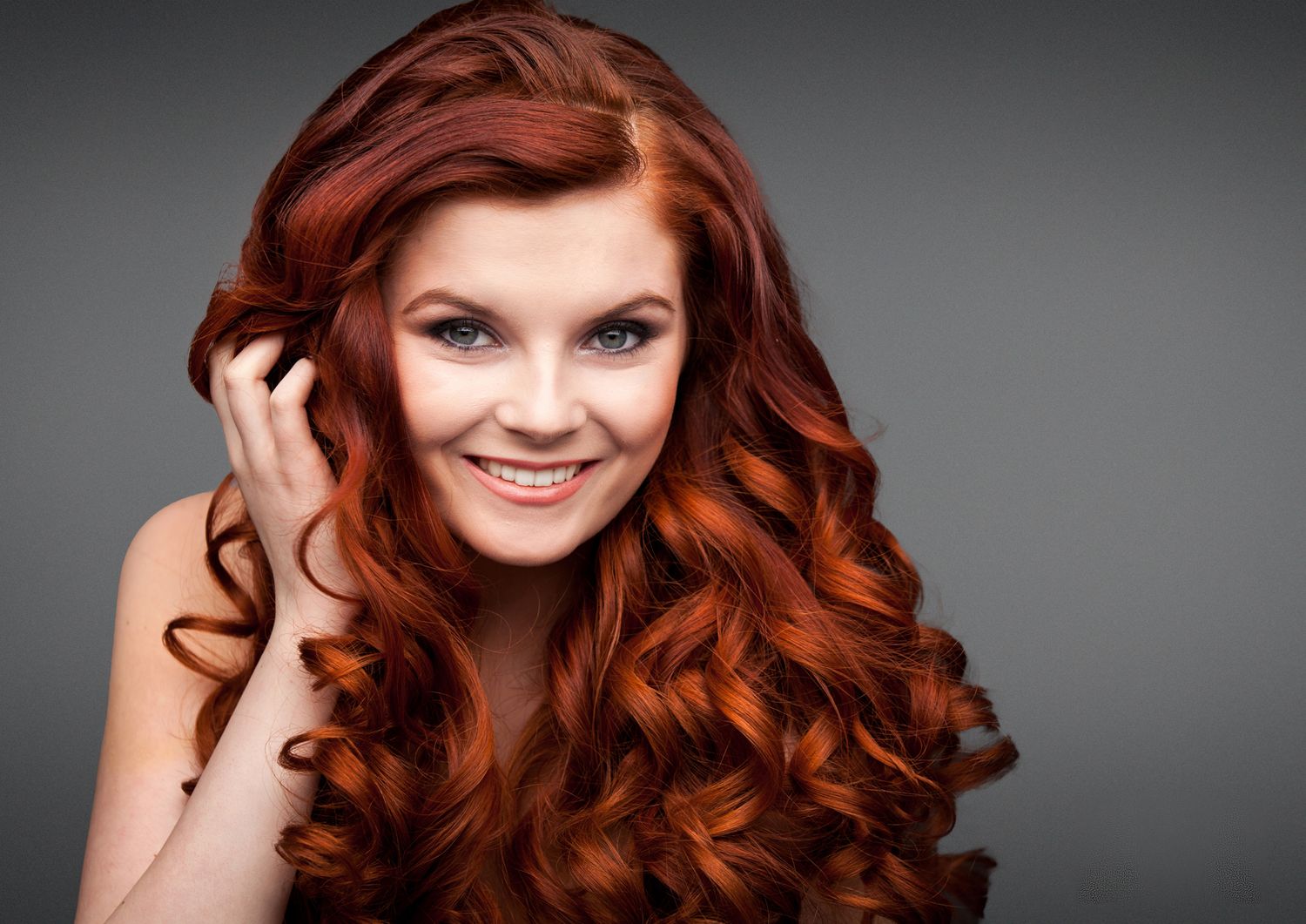 Portrait of a vibrant redhead with long curly hair smiling against a dark background.