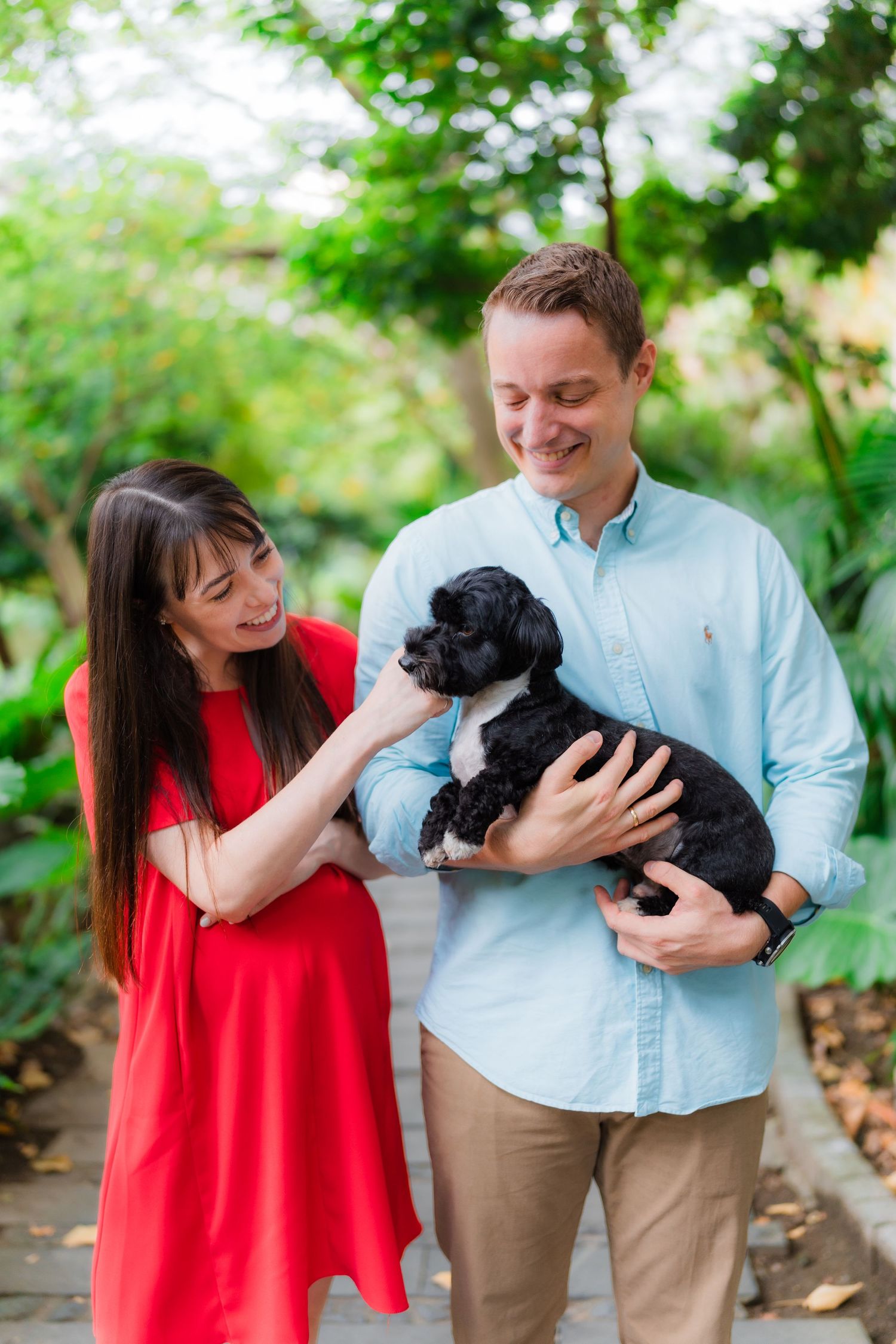 A couple holds their small black puppy while posing together in a garden setting.