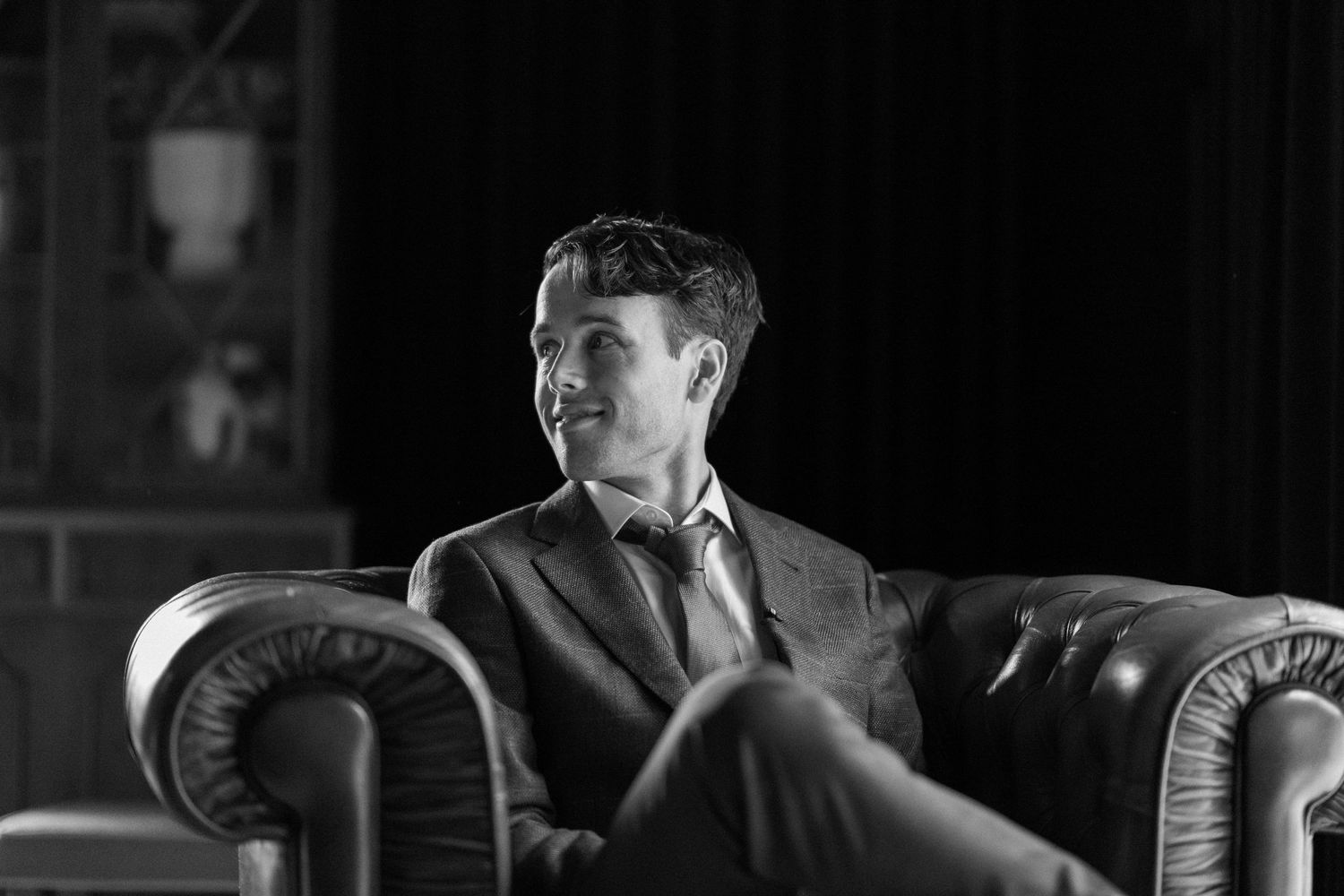 Elegant man in suit sitting thoughtfully in leather armchair in dramatic black and white lighting.