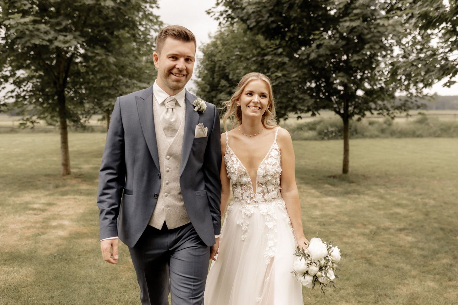 Smiling bride and groom walking hand in hand through a lush green park on their wedding day.