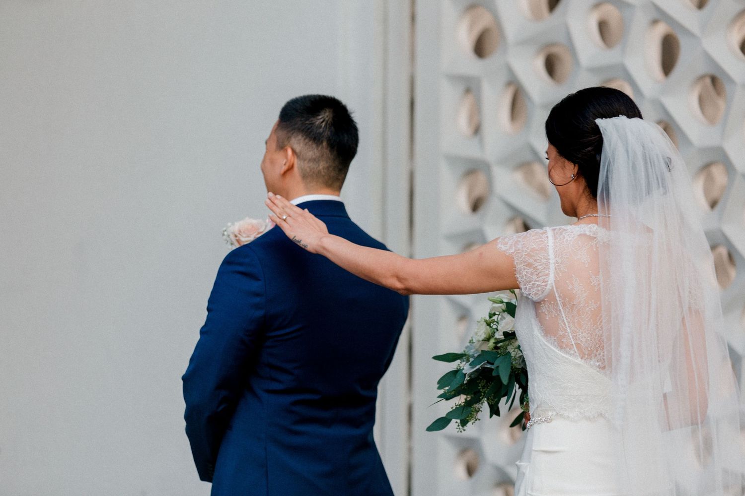 A bride in white follows her partner while holding a green bouquet against a latticed wall.