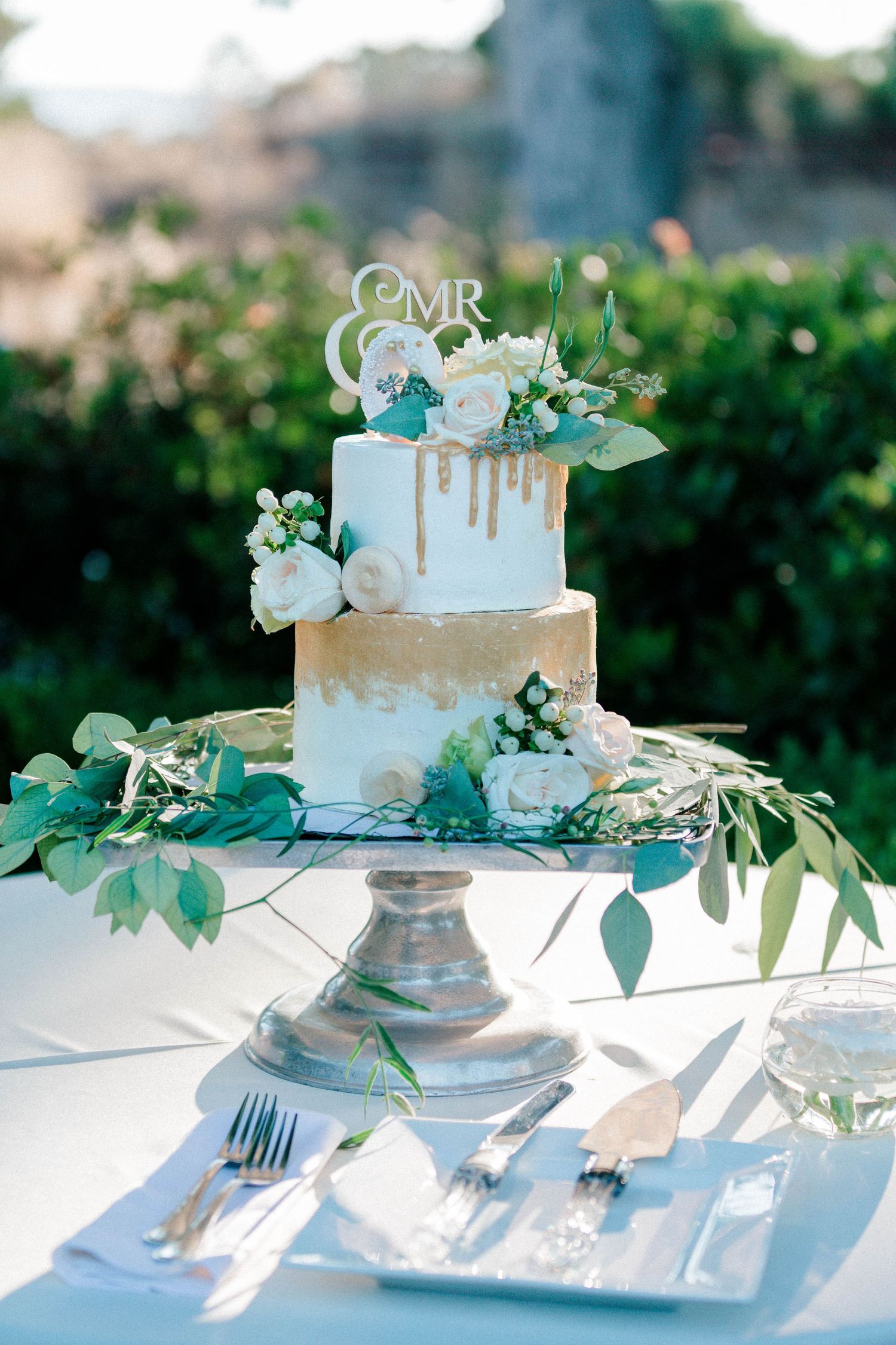 Elegant blue and white wedding cake decorated with gold accents and fresh greenery on a silver stand.