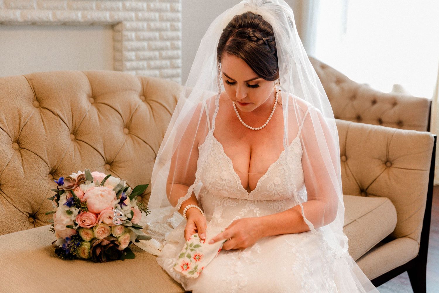 A bride in white lace gown sits on beige couch with floral bouquet nearby.