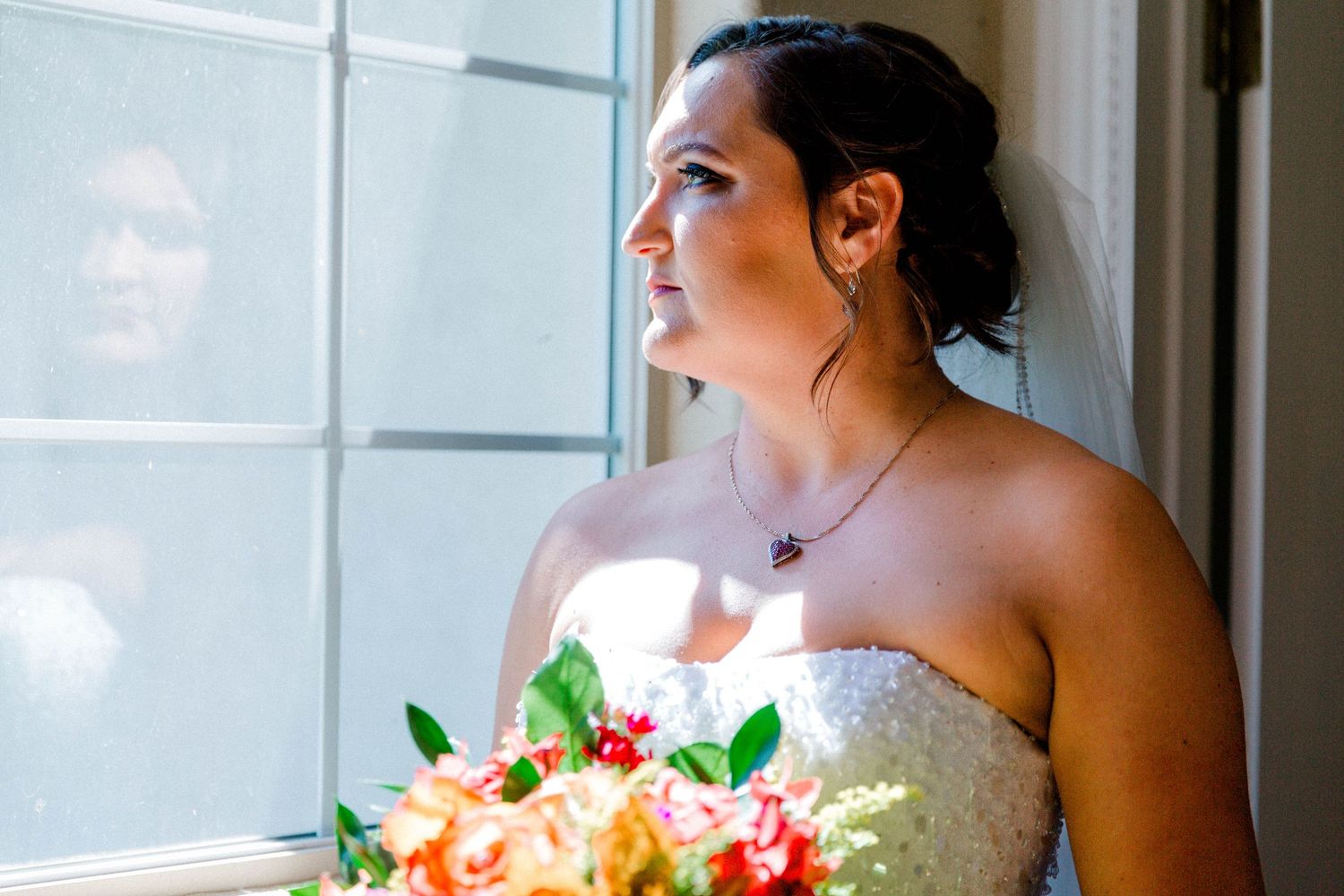 A bride in a strapless gown gazes out a window with soft natural light and peachy floral bouquet.