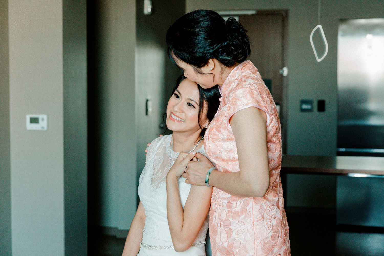 Two figures embrace in soft pink and white dresses against a modern interior background.