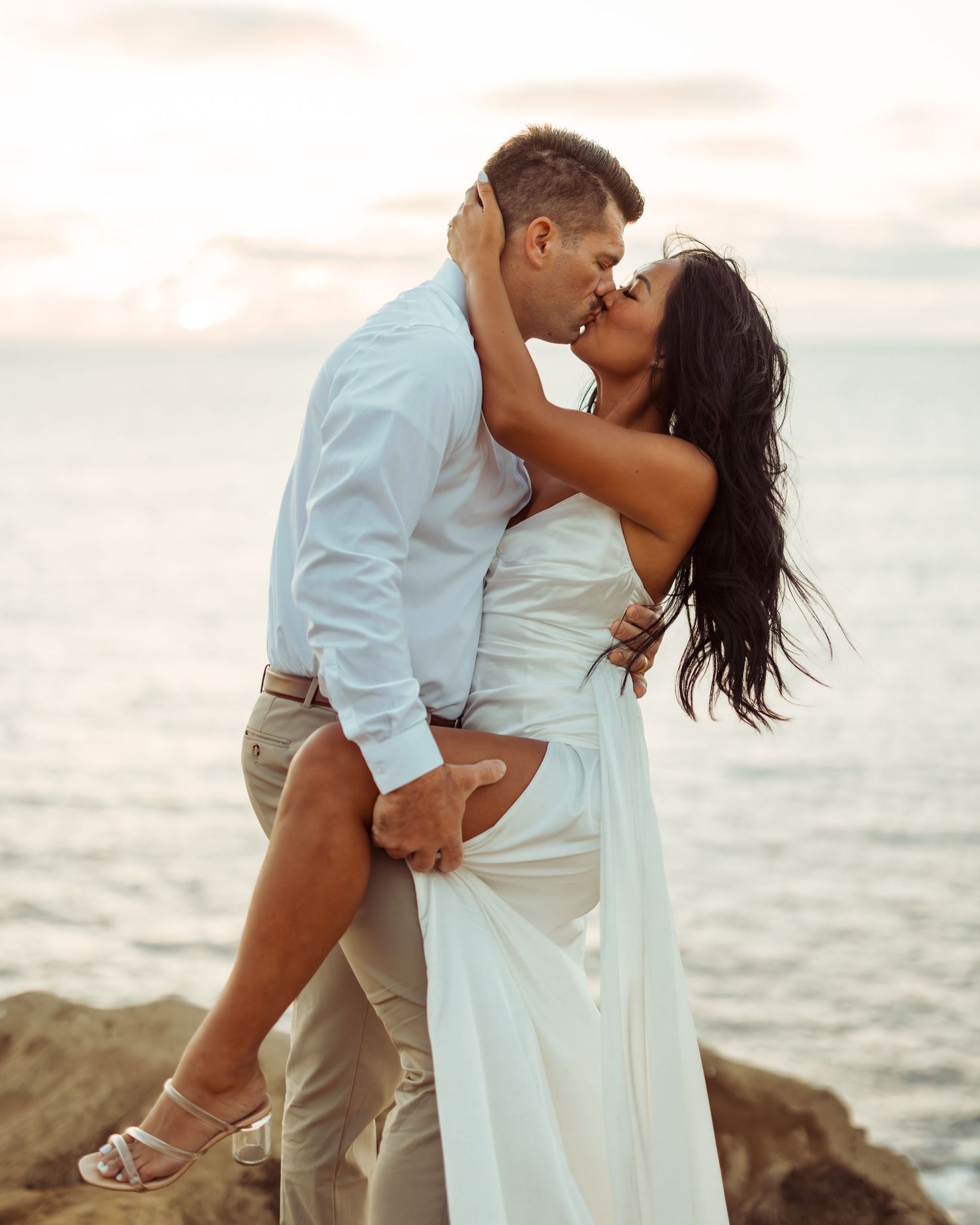 A passionate embrace between a couple in white and beige attire by the ocean.