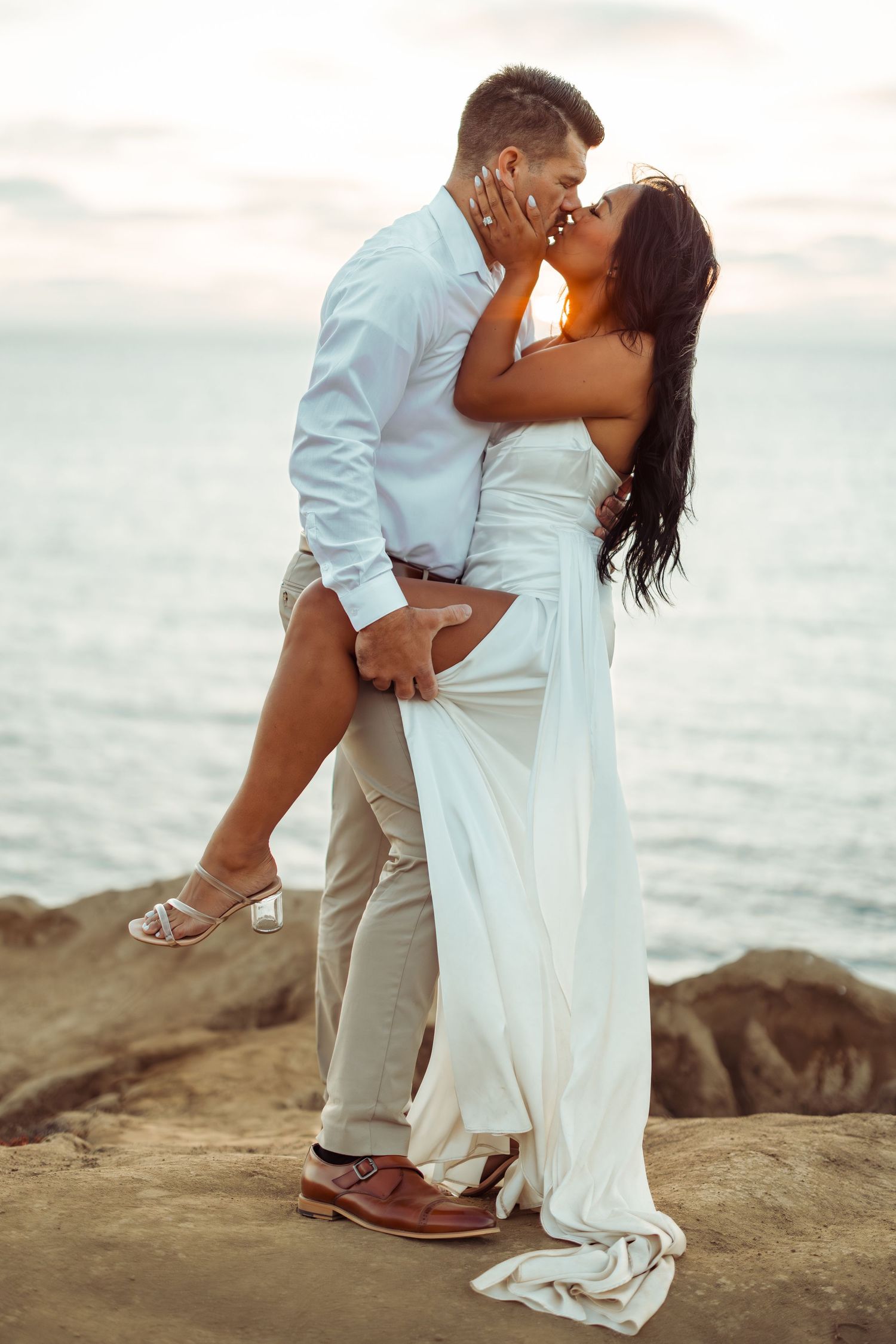 Romantic couple embraces on coastal rocks during sunset as wind blows through white flowing dress.