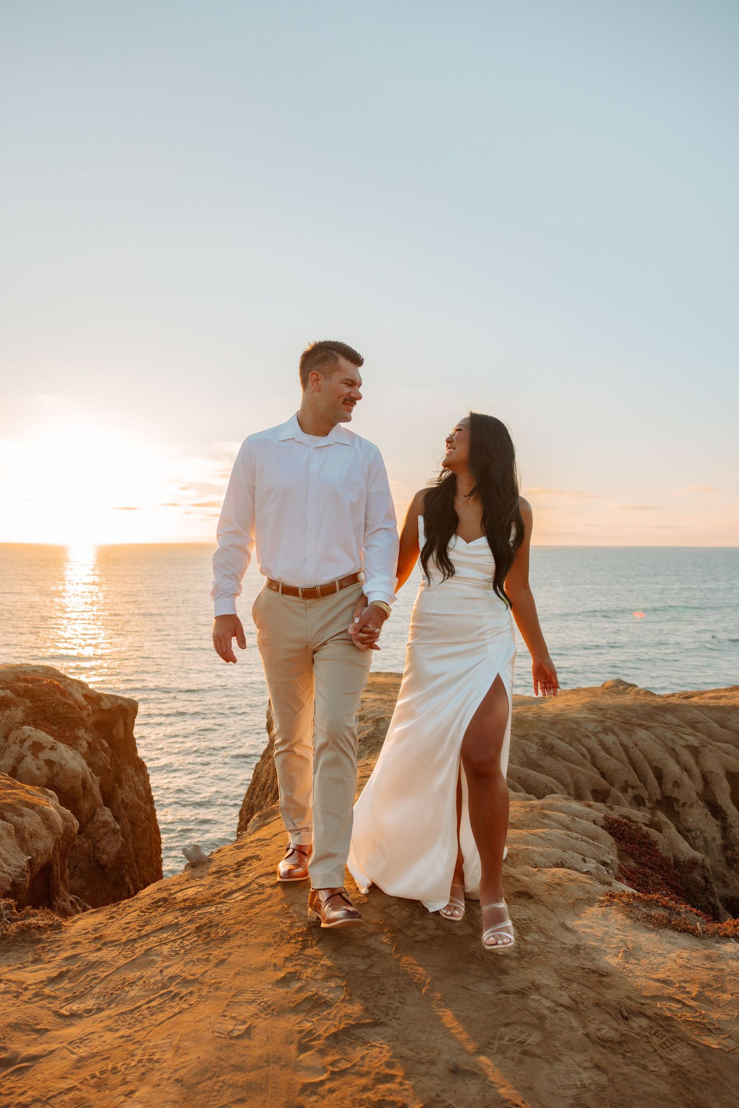 Couple walking hand in hand along rocky coastal path at golden hour.