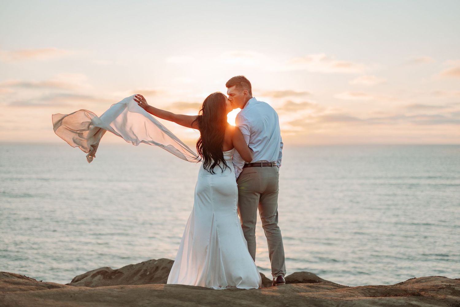 Couple in formal attire dancing together on oceanside cliffs at sunset.