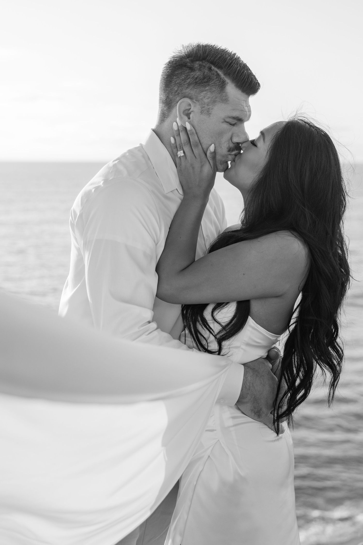 Black and white romantic portrait of couple kissing by the ocean.