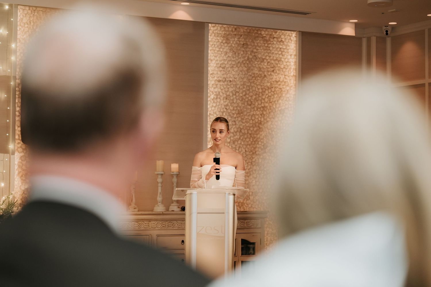 A speaker delivers a wedding speech at a podium with a decorative wall backdrop.