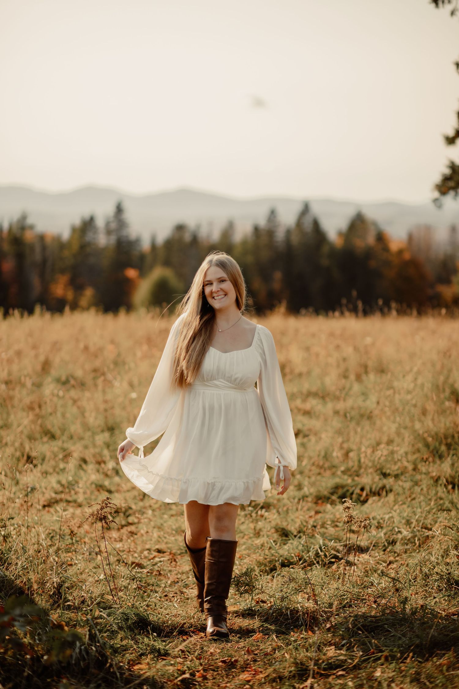Woman in white dress walks through autumn field at golden hour with mountains in background.