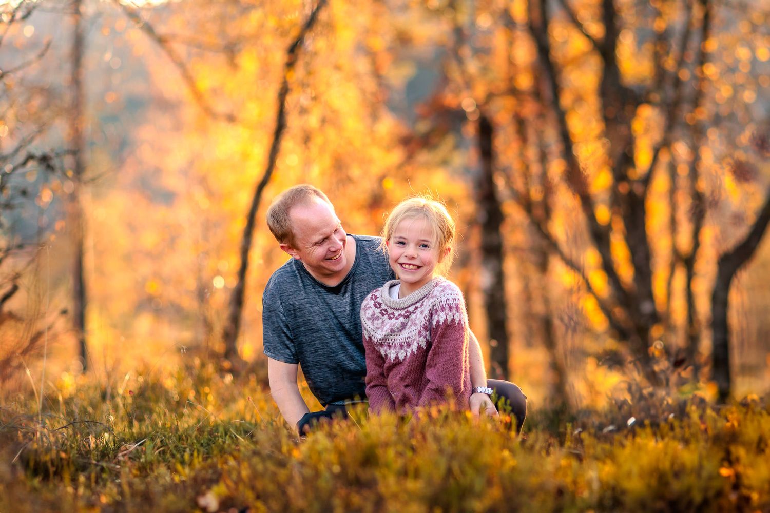 Far og datter sitter sammen i en skog i solnedgang. Faren smiler og ser på datteren, mens hun smiler til kamera.