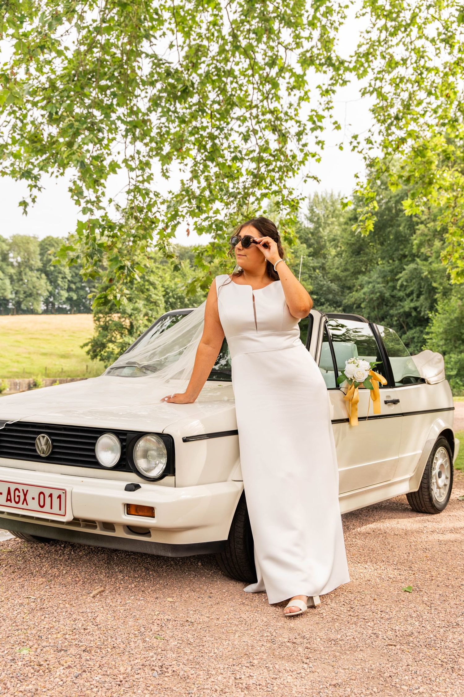 A bride in a white dress poses next to a vintage white convertible car outdoors.