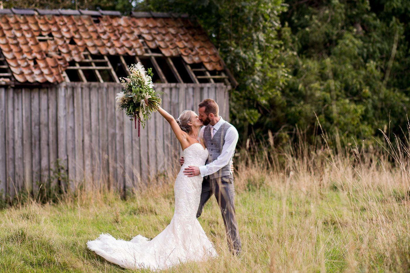 A romantic wedding couple celebrates in a rustic field with an old barn in the background.