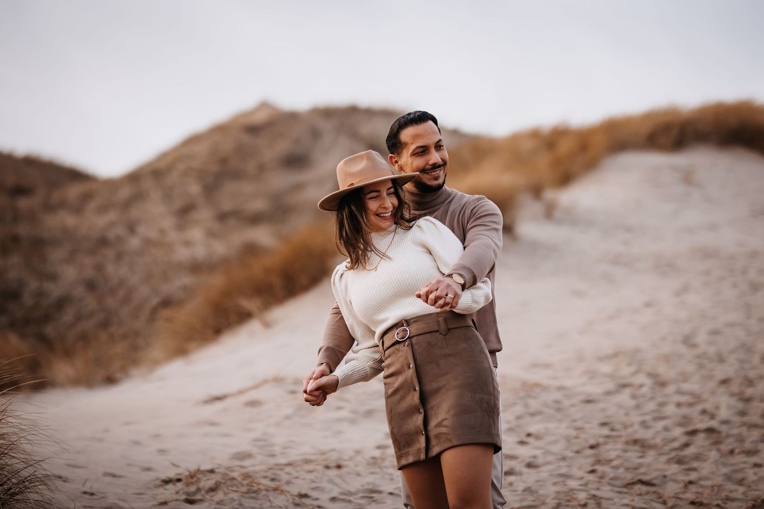Couple embracing and laughing together on sandy dunes during golden hour.