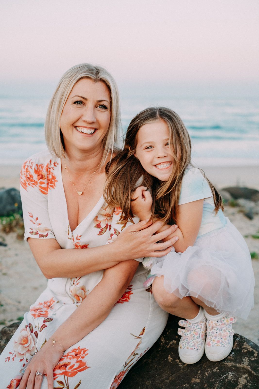 Mother and daughter in matching floral dresses sharing tender moments on coastal rocks.