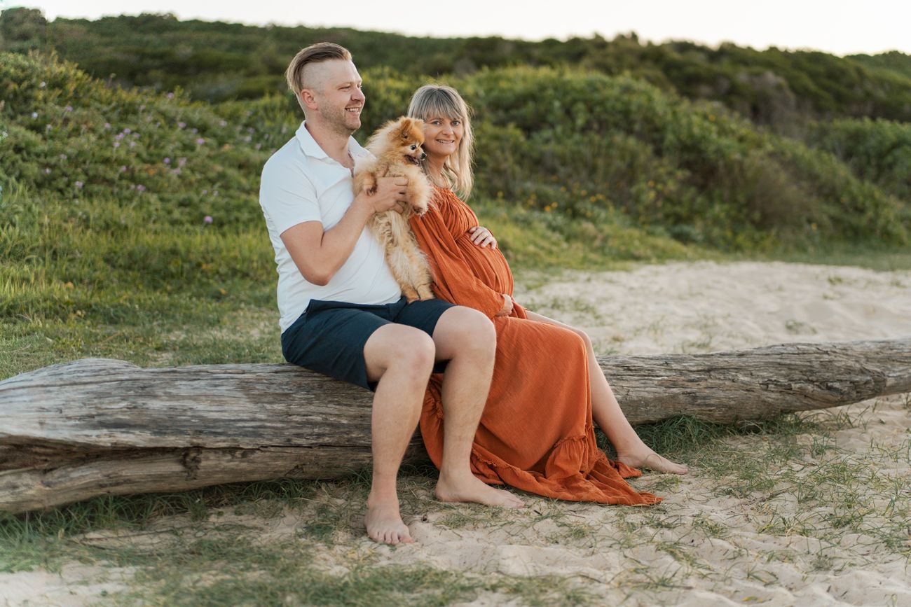 A couple sits together on a fallen log at sunset, with green hills in the background and sand beneath them during a maternity photoshoot