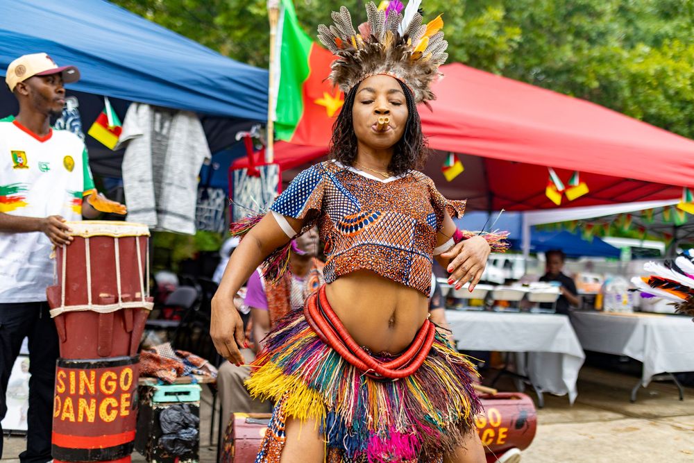 Colorful performers in traditional costumes dance and celebrate at an outdoor cultural festival with tents in background.