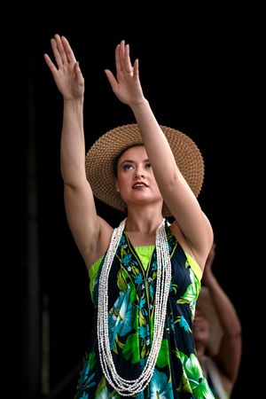 Dancer in a tropical print dress and straw hat performs with arms raised against dark background.