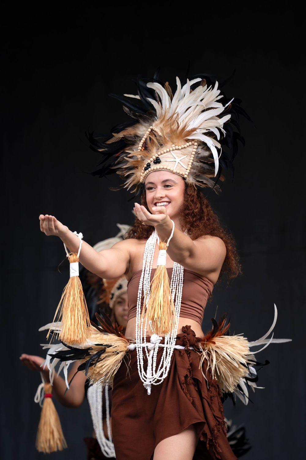 Performer in traditional Polynesian costume with feather headdress dances against dark background.