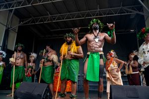 Performers in green aprons and costumes dance on stage during a theatrical production.