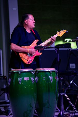 Guitarist performs with electric guitar next to green congas under stage lights.