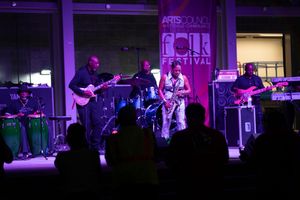 Musicians perform on stage at a music festival with purple and blue lighting illuminating the scene.