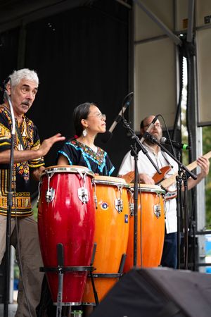 Musicians play a line of red and orange conga drums during an outdoor performance.