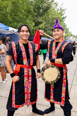 Two performers in traditional Asian festival costumes with red and black garments and decorative sashes pose at an outdoor event.