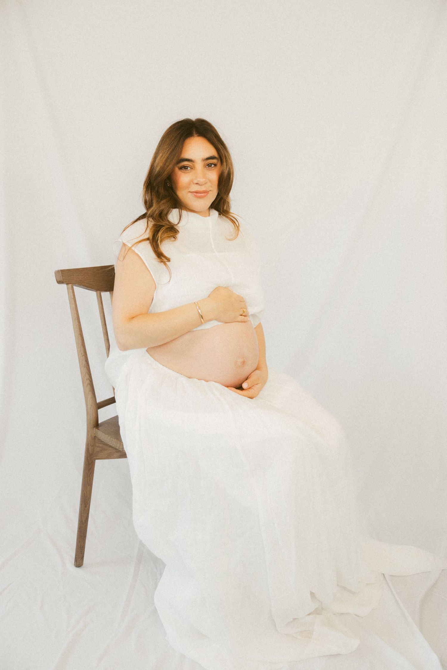 A person in a white flowy dress sits gracefully on a wooden chair against a light backdrop in a minimalist portrait series.