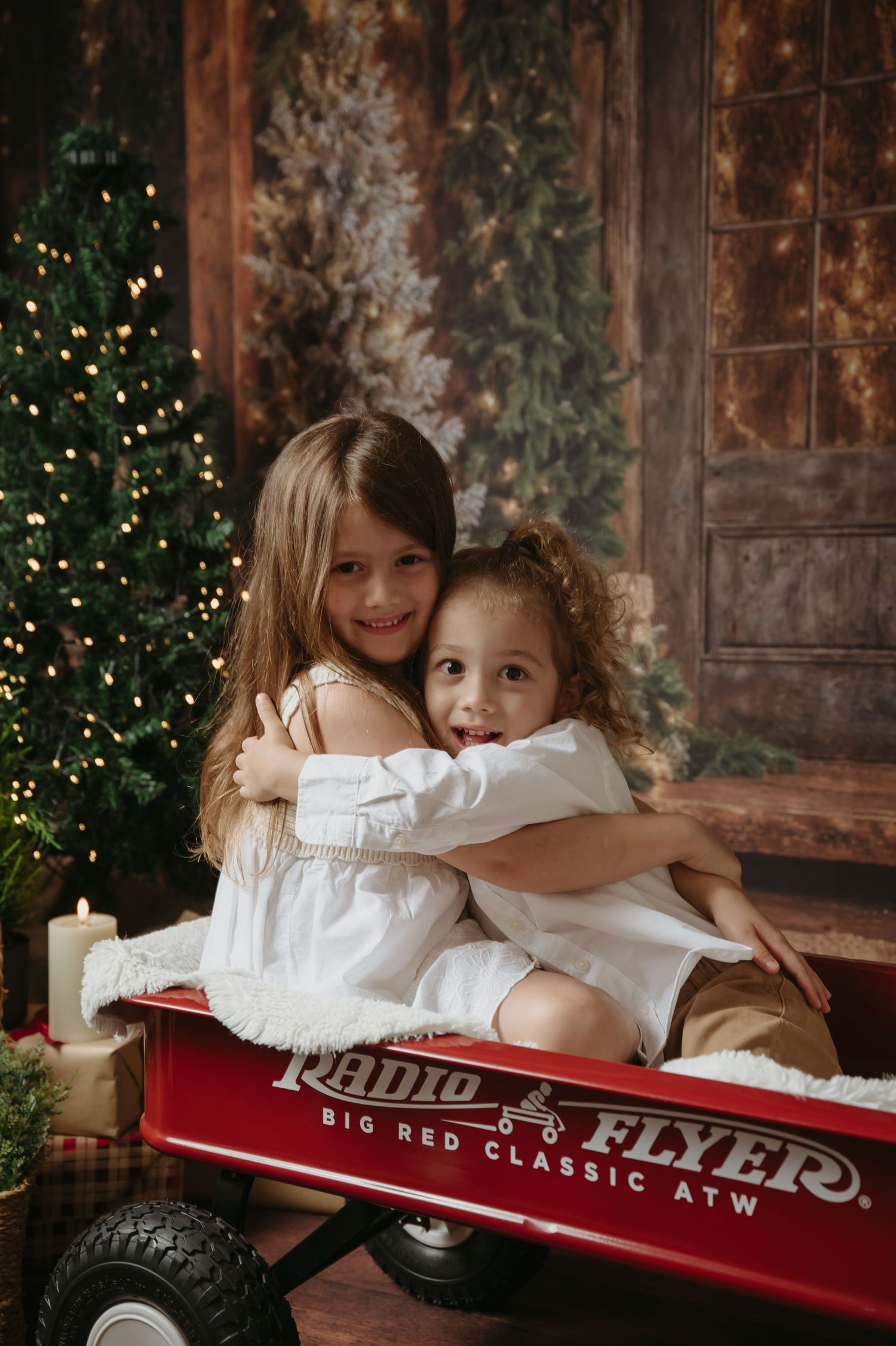 Children playing together in a rustic holiday setting with Christmas trees and vintage red wagon decorations.