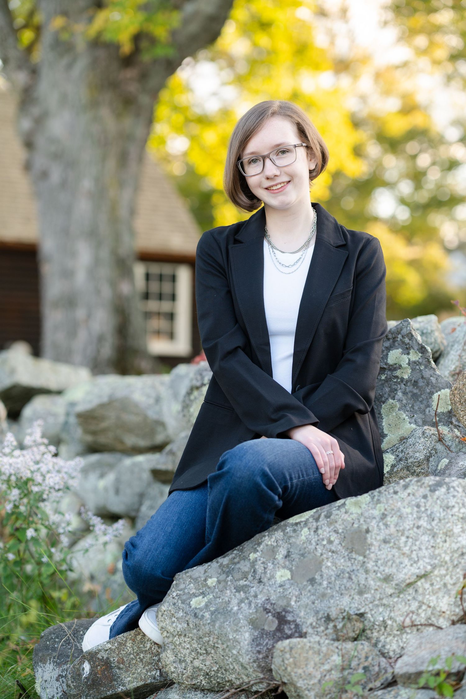 Professional outdoor portrait session showing someone in a black blazer and jeans sitting casually on large rocks in autumn light.