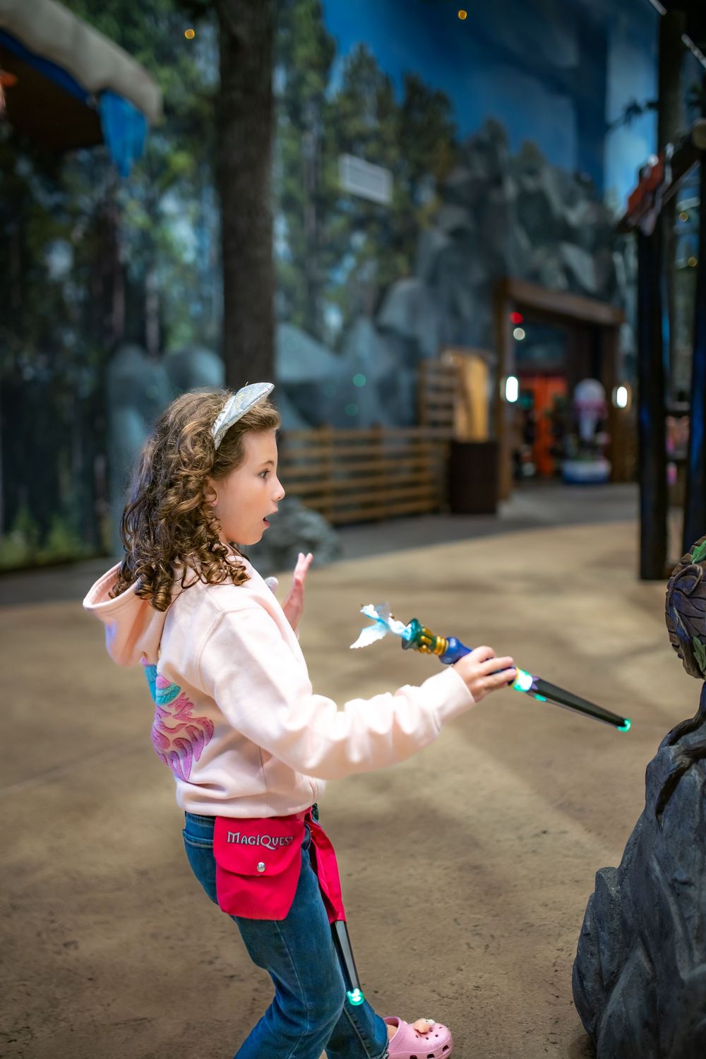 A young child in a pink sweater and red bag runs excitedly through a faux wooded path.