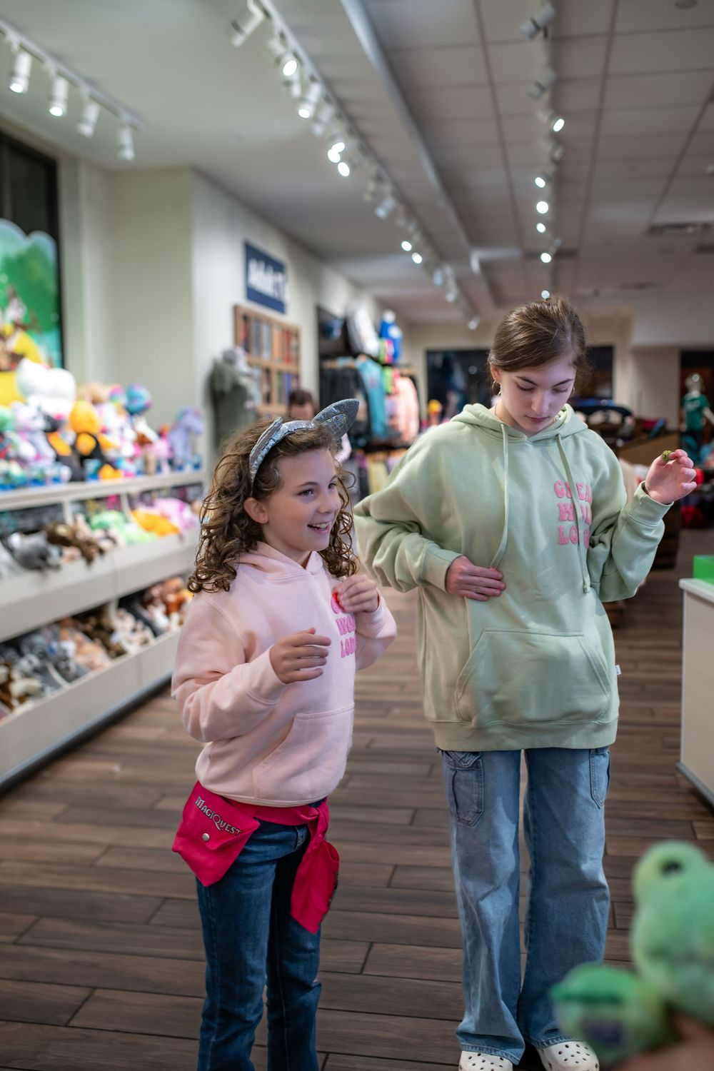 Two young shoppers in hooded sweatshirts browse merchandise at a retail store with stuffed animals and toys on display.