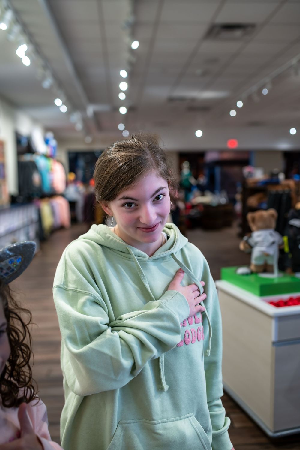 A person in a mint green hoodie sweatshirt poses playfully in a retail store setting with overhead lighting.