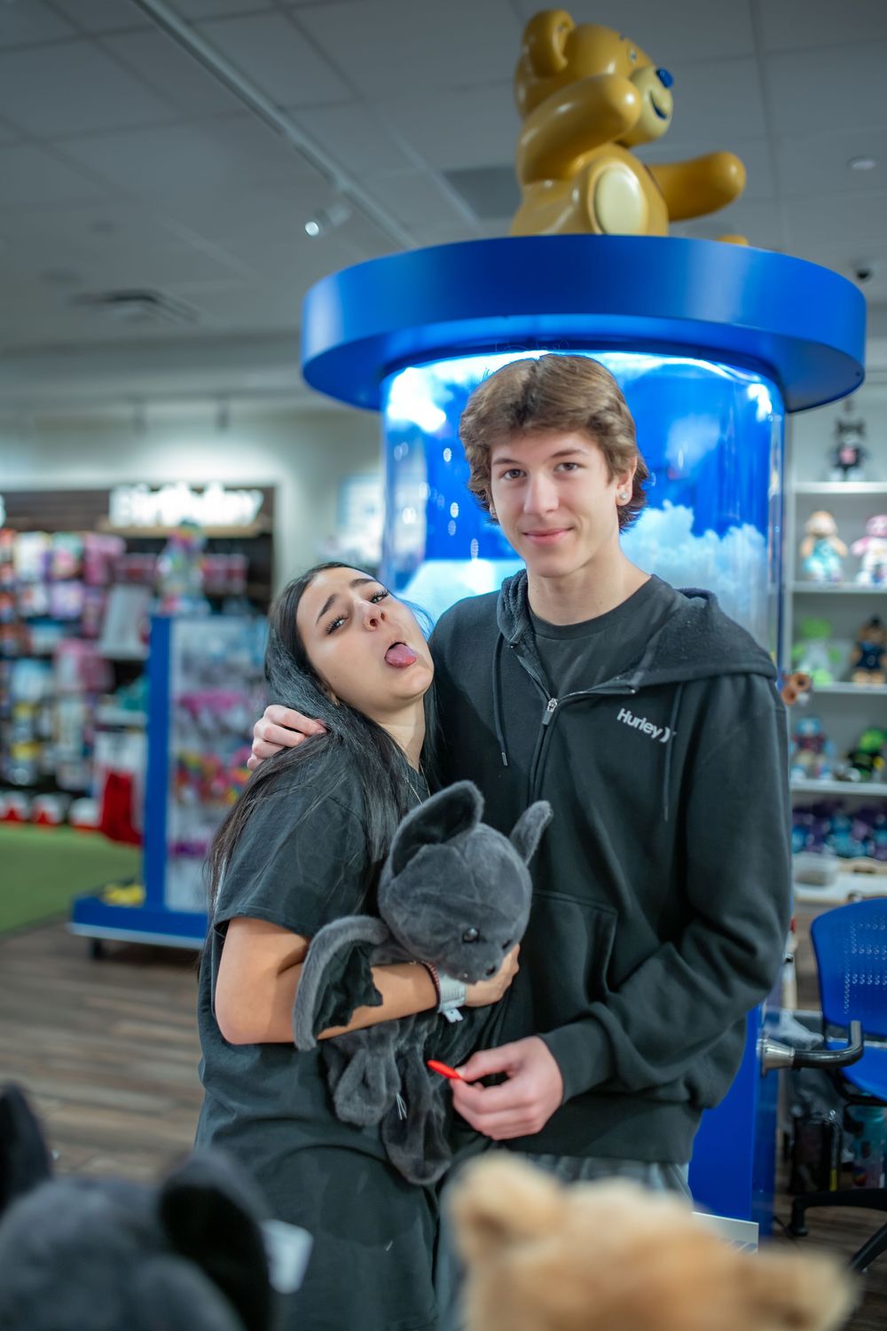 A couple poses with a small gray bat in a Build-A-Bear store with blue display fixtures in the background.