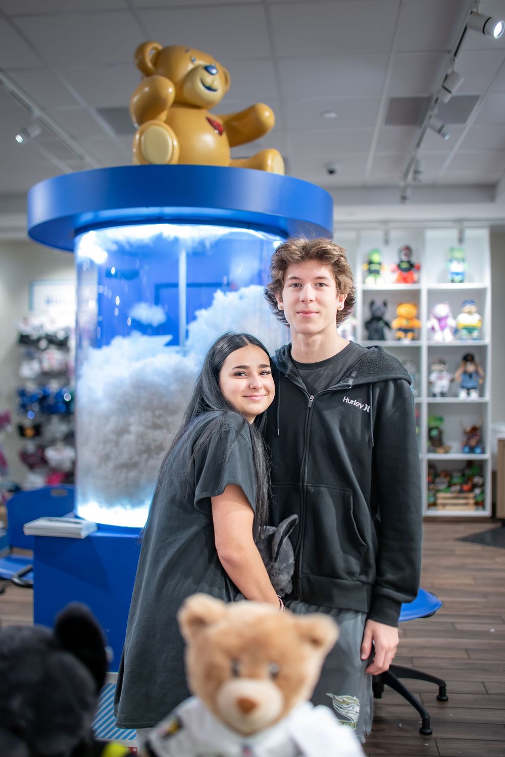 Two people stand in front of a blue display with a large teddy bear decoration at a retail store.