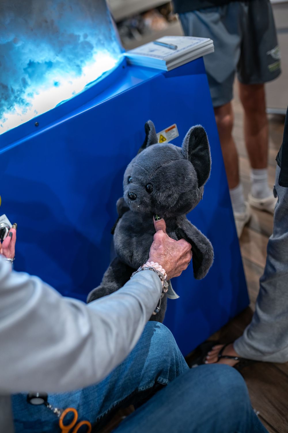 A small gray plush toy being held out against a bright blue background with people standing nearby.